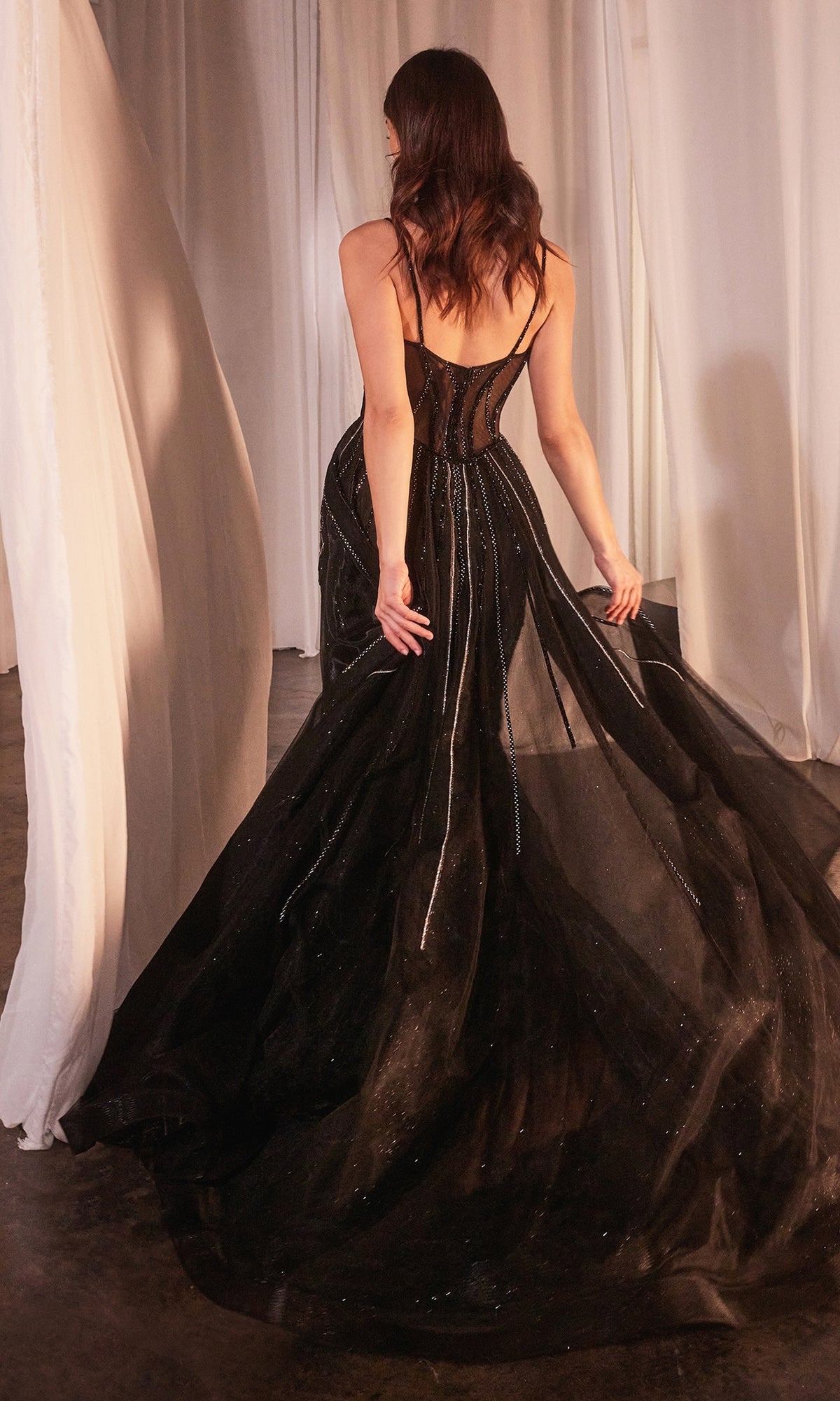 A woman with wavy brown hair stands among white curtains, holding the flowing skirt of the Beaded Overskirt Long Formal Prom Dress CD863—a sheer black gown with sparkling details reminiscent of a formal prom dress.